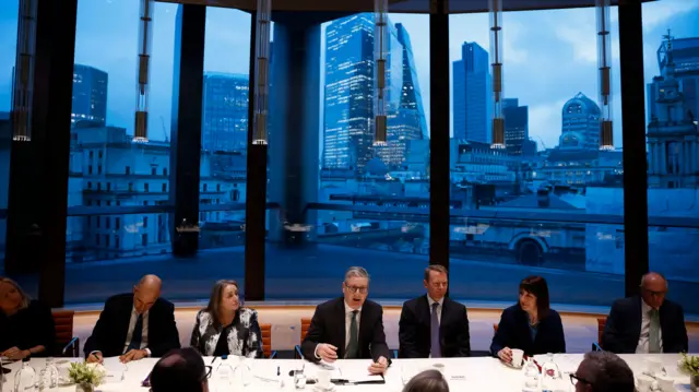 Prime Minister Keir Starmer and Chancellor Rachel Reeves sit among a row of business leaders during a meeting, with a giant glass window in the background overlooking high-rise buildings.