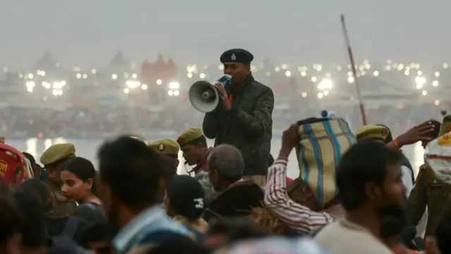 An officer in uniform speaks into a megaphone as he stands above a crowd of people. In the background is a river and a blurred cityscape.