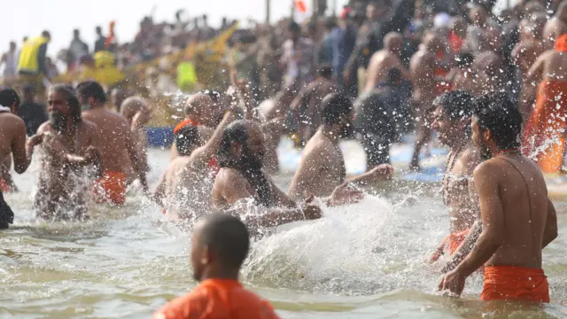 A group of men, some wearing orange shorts and tops, bathe in the river with water splashing