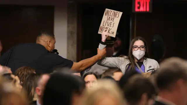 A man tries to remove a protestor wearing a health face mask and holding up a sign that says "Vaccines save lives. No RFK Jr" during the confirmation hearing in Washington.