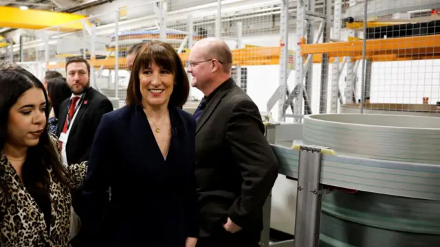 Rachel Reeves in navy blazer walking past other people in a warehouse, smiling