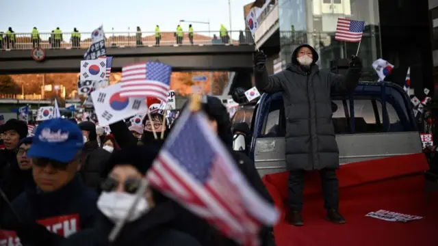 Front view of protesters holding South Korean and US flags on the road, with a man standing on the back of a truck holding a US flag