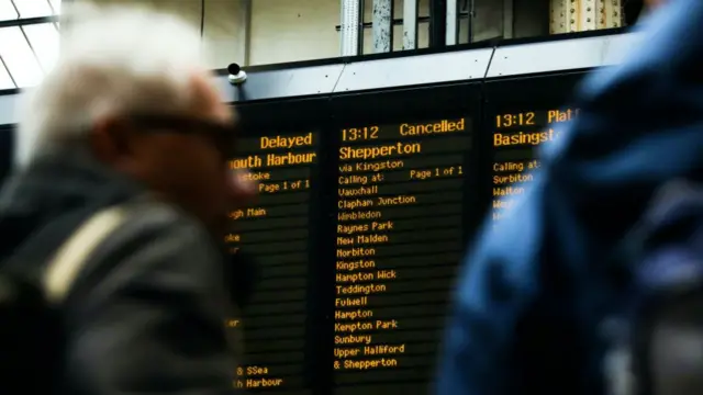 Blurred passengers observe train board at station. Several services are marked as delayed or cancelled