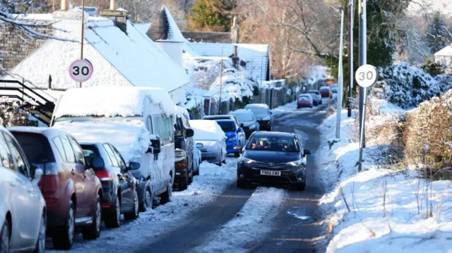 A car drives through snow in Balerno, Edinburgh. Nearby houses to the left of the frame are covered in snow as are the cars parked to the side of the road