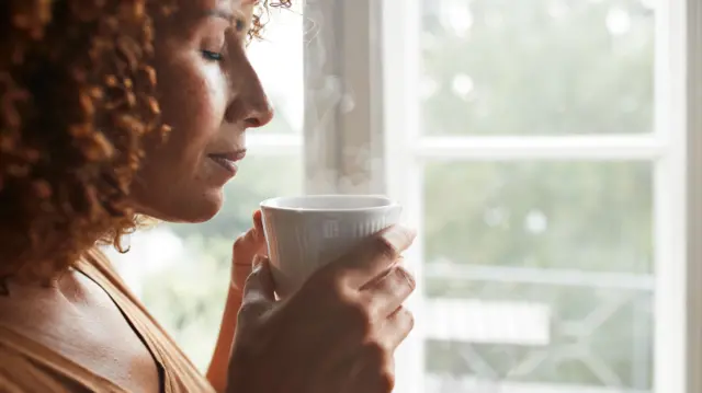 A woman holds a white cup close to here lips, with her eyes closed. Steam curls off the cup.