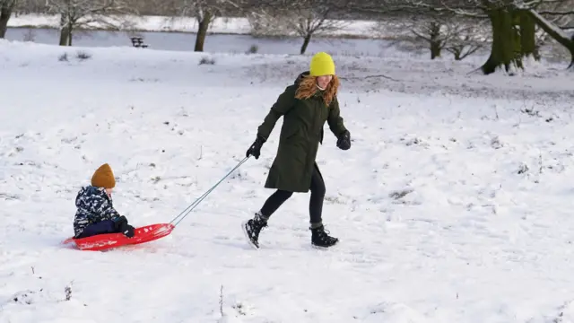 A woman in winter wear with a bright yellow woollen hat drags a child in winter jacket and light brown hat across a snowy park in a red sledge. A body of water with snowy banks and tree trunks are in the background