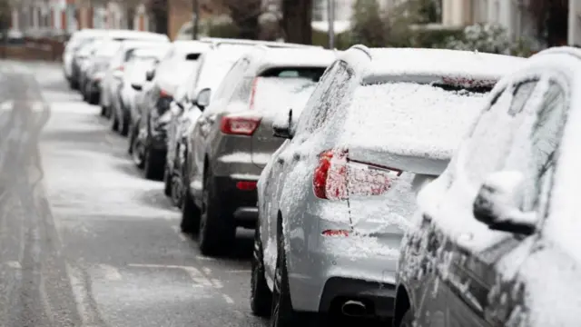 Parked cars covered in thin layer of snow. Bottom part of a row of terraced houses visible from the top right to the top left of the image