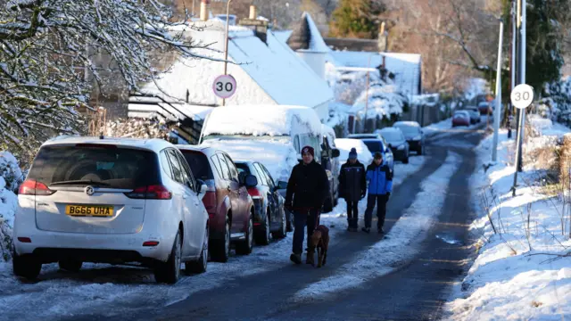 People walk through snow in Balerno, Edinburgh, past cars covered in snow at the side of the road