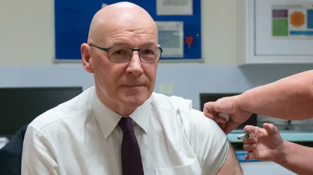 John Swinney with a nurse injecting his flu vaccine. He's sitting down with his left shirt sleeve lifted as a nurse administers the vacchine. Behind him a blue board with a poppy and two sheets of paper with graphics and medical equipment including tables