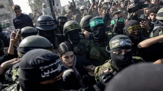 Arbel Yehud surrounded by armed militants with their faces covered and a large crowd in the background.