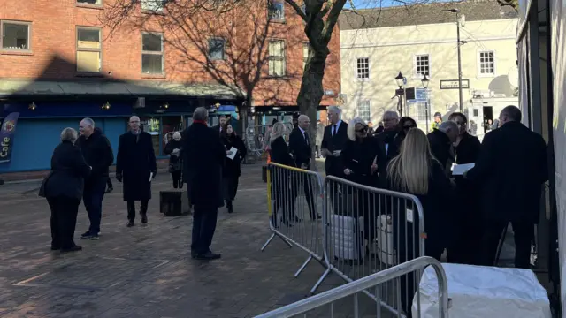 A small crowd of people, dressed in black, mill about in the paved area outside Hull Minster