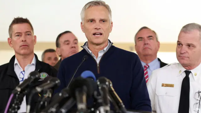 CEO of the American Airlines Robert Isom addresses the media. He is stood in front of a podium that has lots of black microphones attached to it. He is wearing a navy jumper, and he has grey hair and blue eyes.