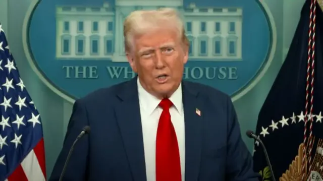 Donald Trump mid-shot at lectern delivering speech in dark suite with white shirt and red tie