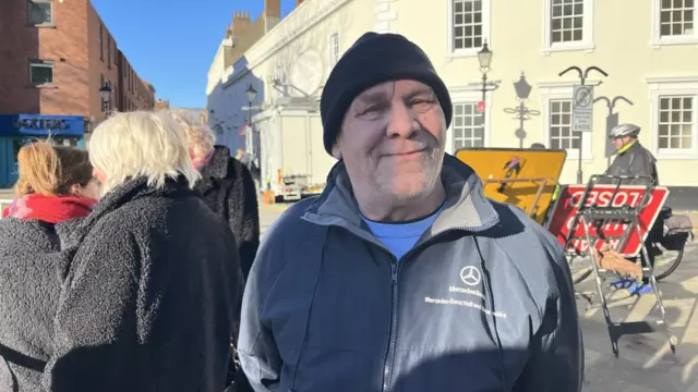 A man in a black beanie and Mercedes-branded blue jacket smiles at the camera. A cyclist can be seen riding to his left, while a group of three women in black coats stands to his right