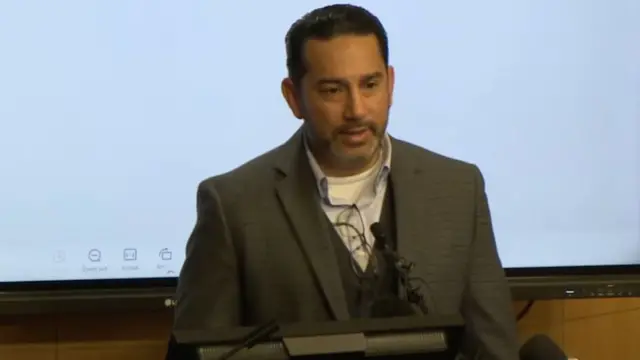 Jesse Romo stands at a podium to address the media. He is wearing a grey blazer and waistcoat, and he has black hair.