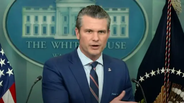 Pete Hegseth mid shot at podium standing up in dark blue suit, striped tie (blue, red and white) and striped blue shirt