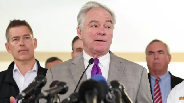 US Senator Tim Kaine addresses the media at a podium that has lots of black microphones attached to it. He is wearing a grey blazer, white shirt, and purple tie, and he has white hair and blue eyes.