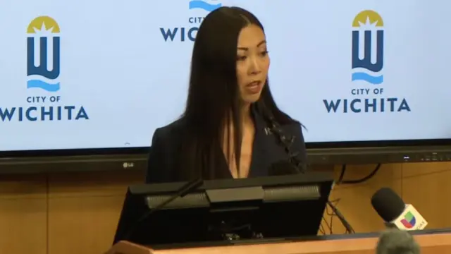 Wichita Mayor Lily Wu stands at a wooden podium to address the media. She has long black hair and is wearing a black blazer.