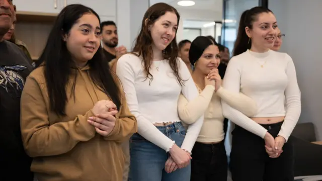 Four young women stand in a line, looking past the camera with emotional expressions on their faces