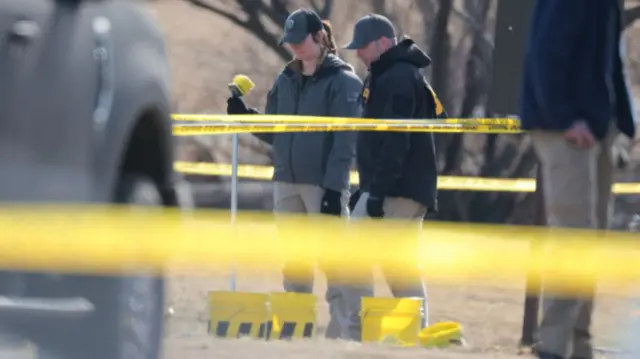 Two people stand looking over a bright yellow bucket and they are wearing baseball caps. There is yellow tape surrounding the area around them.