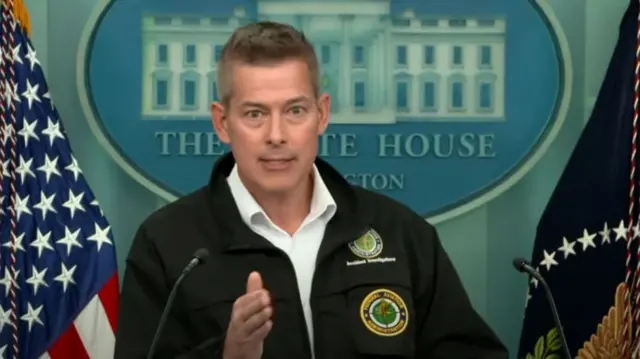 Sean Duffy mid shot standing up while addressing press. His right hand is partially extended in front of him, he's wearing a black jacket with the Department of Transportation logo to the top left pocket and lapel