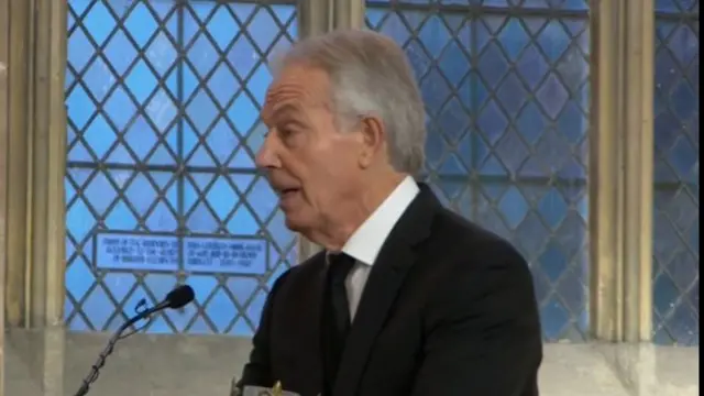 Tony Blair stands in black suit, black tie and white shirt as he delivers eulogy. Church window behind him