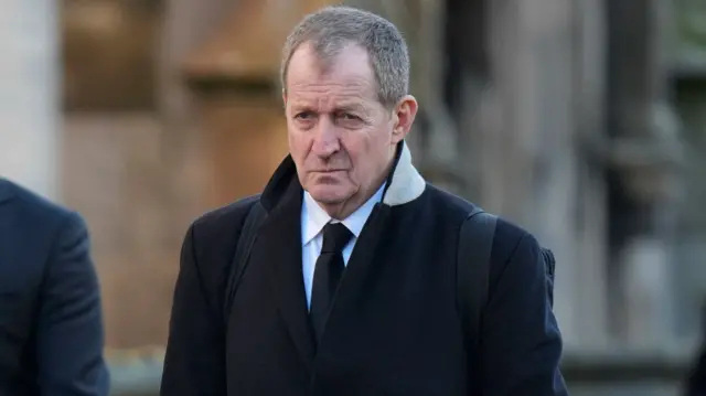 Alastair Campbell, wearing smart black clothing, walks as he approaches the ceremony outside