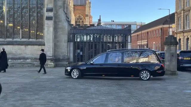 Hearse driving towards the entrance of Hull Minster, a member of the funeral home walking in front of the behicle