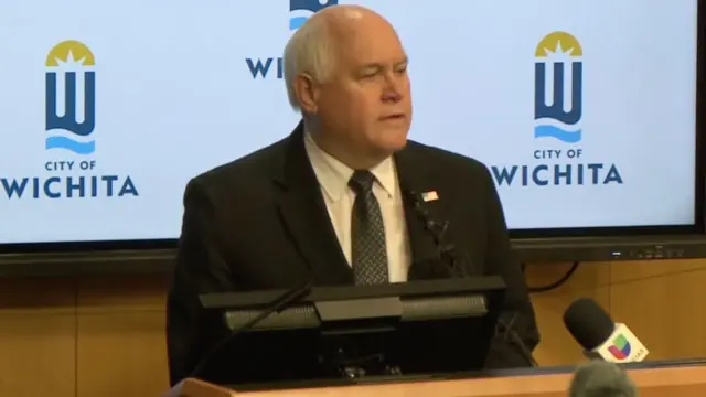 Ros Estes stands at a wooden podium to address the media. He is wearing a black blazer, white shirt, and a black and grey patterned tie.