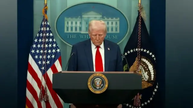 Donald Trump in dark blue suit, red tie and white shirt with his head bowed, US flag to his right, dark blue flag with presidential insignia to his left