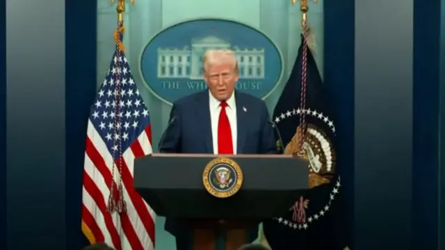 Donald Trump addresses press from White House. He's standing behind a lectern in a dark blue suit with red tie and white shirt. A US flag to his right and dark blue flag with the Presidential insignia to his left