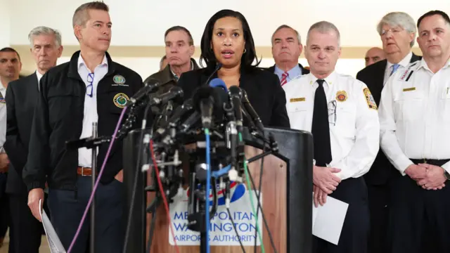 Washington DC Mayor Muriel Bowser addresses the media. She is stood at a podium with many microphones attached to it. Men, who are representing different authorities, are stood behind her.