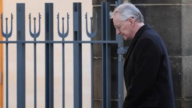 Benn, in a black coat looks down as he walks to the left