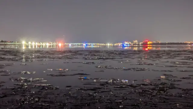 Ice floats on the Potomac River. Red and blue emergency lights can be seen in the distance.