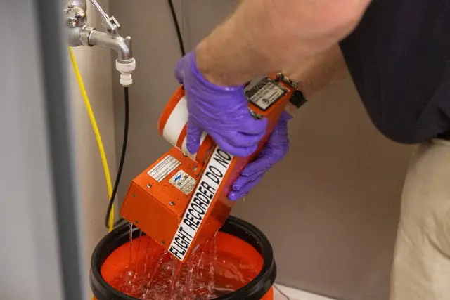 A person wearing purple rubber gloves holds a flight recorder, and pours water from it into a bucket