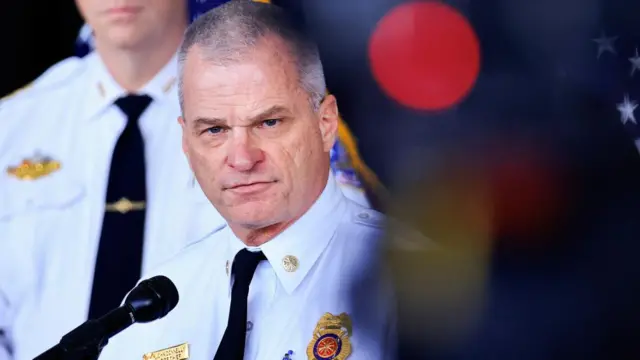 DC Fire Chief John A. Donnelly speaks to the media. He has short, greying hair and blue eyes. He is wearing a white shirt with a black tie and a badge representing his fire department on his chest.