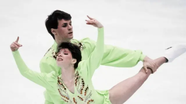 A man and woman skating, in green bedazzled costumes. The woman holds both hands above her head, whilst the man behind her holds one of her legs up