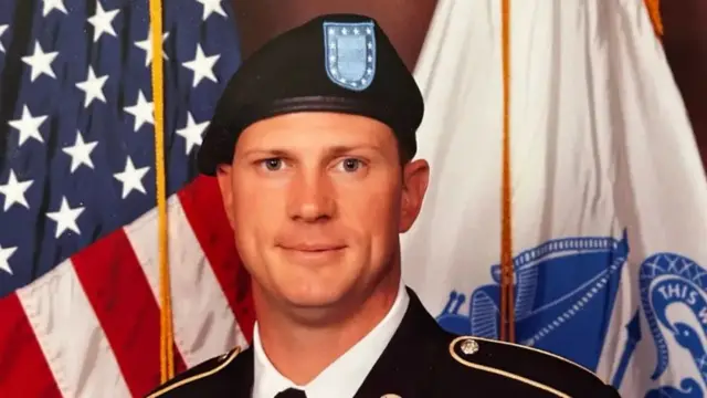 Andrew Eaves in black uniform looking at the camera with flags behind him