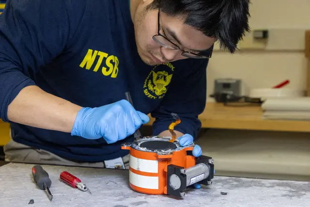 A man wearing a jumper with NTSB working on a small round orange and white device with various hand tools