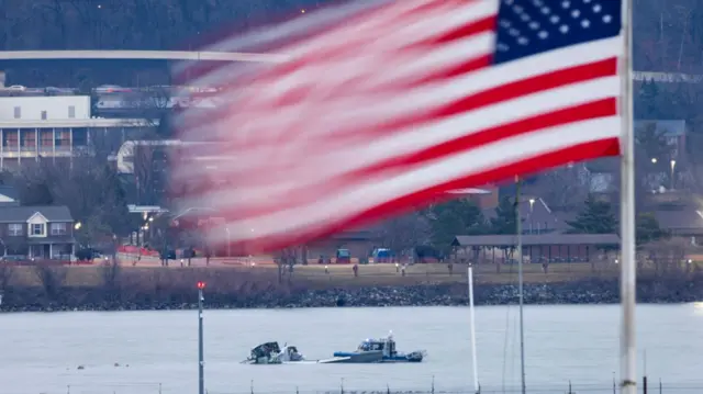 Coast Guard boat guards debris of commercial plane in Potomac River. An American flag, blurred, is flying in the foreground