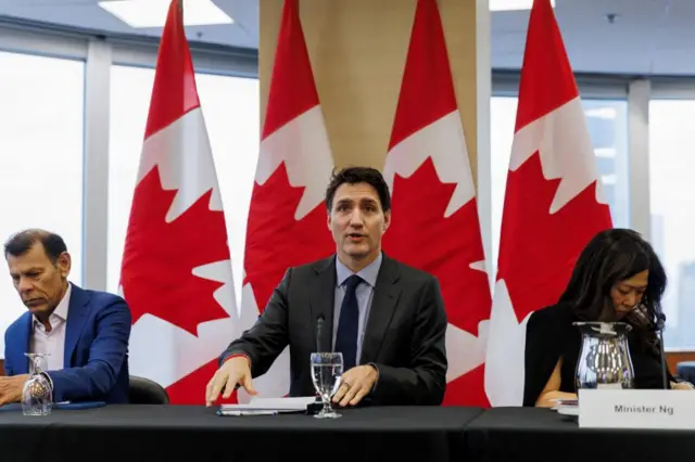 Justin Trudeau sits between two others speaking into a microphone. Canadian flags are draped behind him