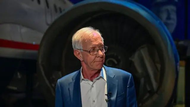 Sully Sullenberger looks to his left. The engine of a plane can be seen in the background.