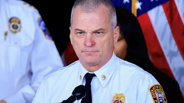DC Fire Chief John Donnelly speaks to the media. He has short greying hair and blue eyes. He is wearing a white shirt with a black tie and his fire department's badge on his chest and sleeve.