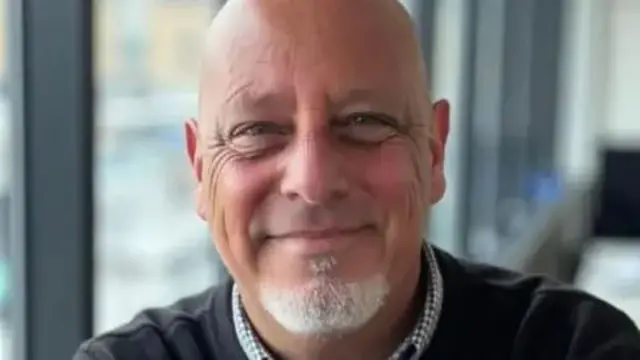 Ian Epstein looking into the camera and smiling, with black jumper and collared shirt and white beard