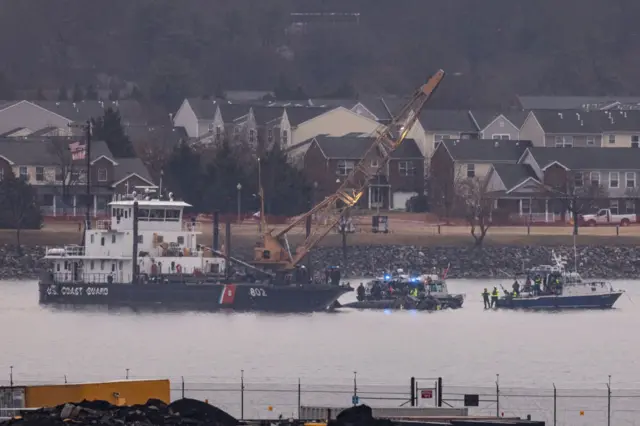 A large rescue boat in the river with a large yellow crane on the front of it. Two smaller boats are also in the water. Several houses are in the background.