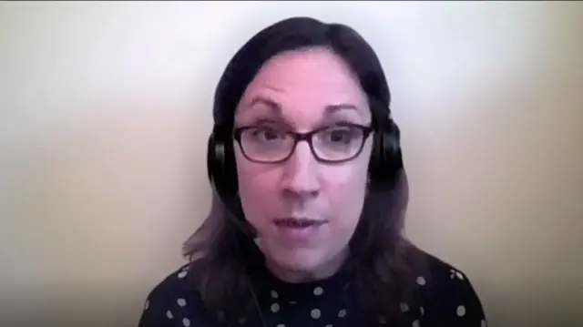 British Red Cross spokesperson (medium brown hair, glasses, polka dot shirt) interviewed via video link. She's wearing headphones with a speaker, a white wall behind her