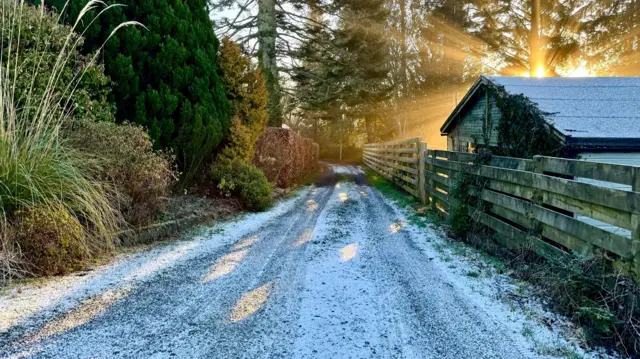 A path dusted in snow is at the centre of the image. A wooden building to the right and trees to the left and ahead. The sun is rising in the right