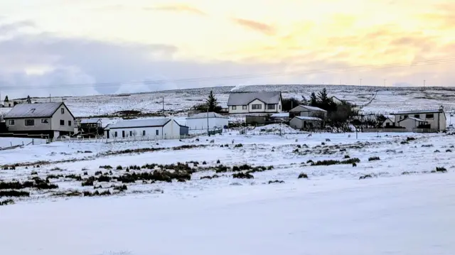 Snow covers the hills with some buildings seen at the centre of the image. The sun rises in the right hand corner giving a yellow/orange sky