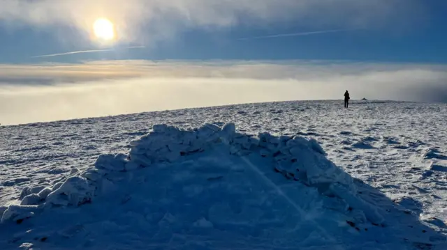 Lone figure on the right side of the photo walks along the top of the snowy mountain, the sun shining to the top left of the image. Sky is blue, with low white clouds towards the top of the image