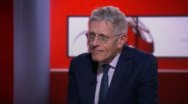 Simon Calder is shown sitting in the BBC News studio. He is dressed in a suit and sits in front of a red background with the BBC news logo.